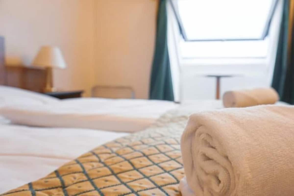 A hotel room with a bed, a rolled towel, a lamp, and a window with curtains.