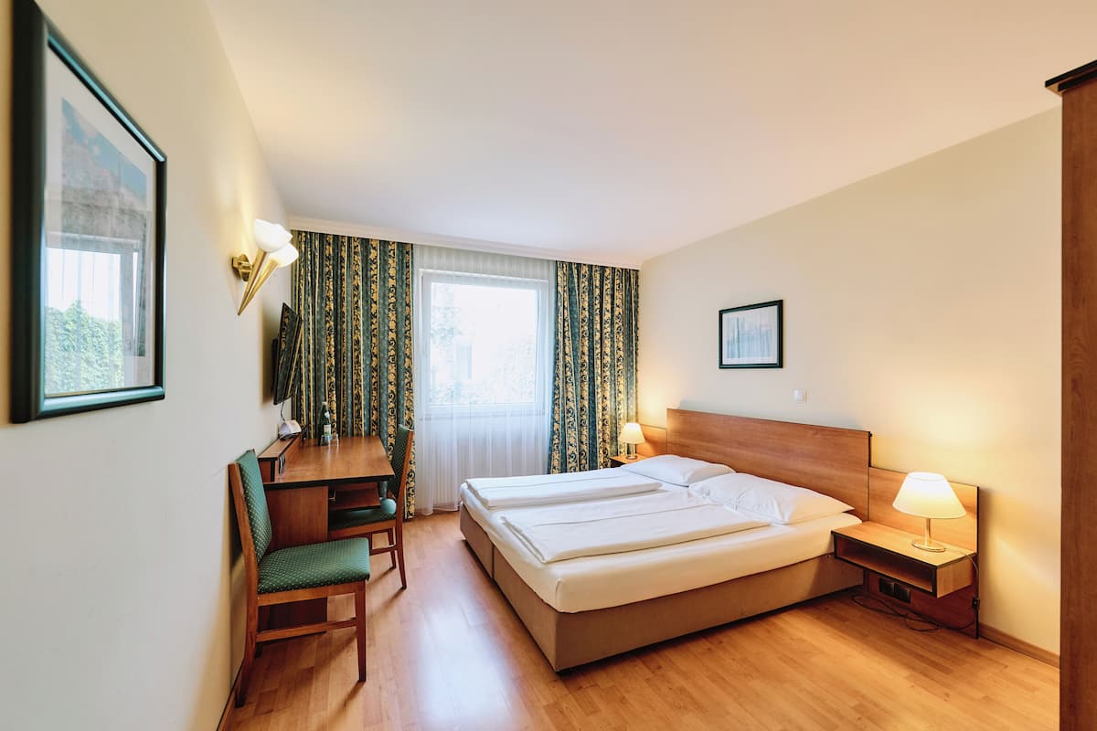 A hotel room with a bed, bedside table with lamp, desk with chair, and curtains.