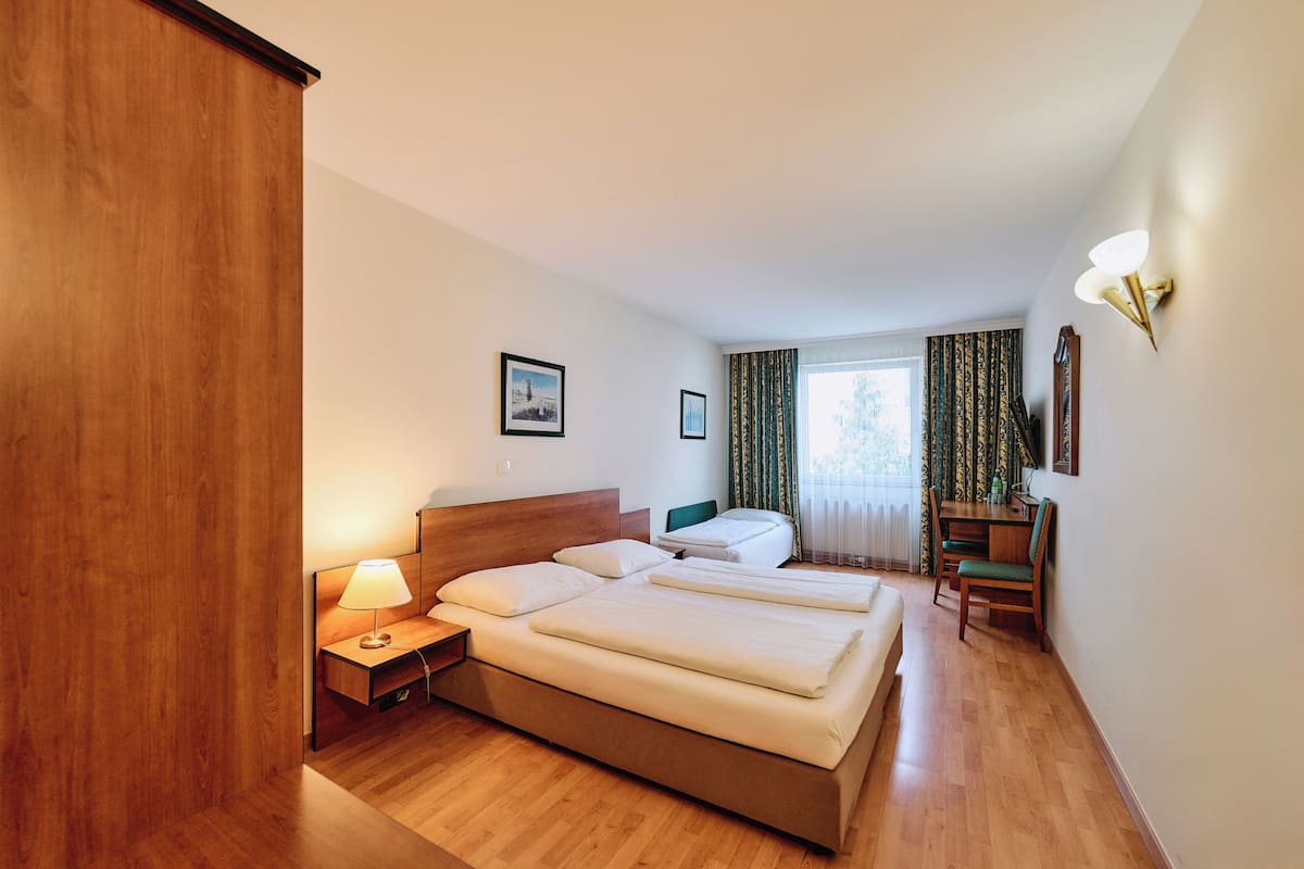 A hotel room with a wooden floor, a bed with two pillows, a bedside table with a lamp, a desk with a chair, and a window with curtains.