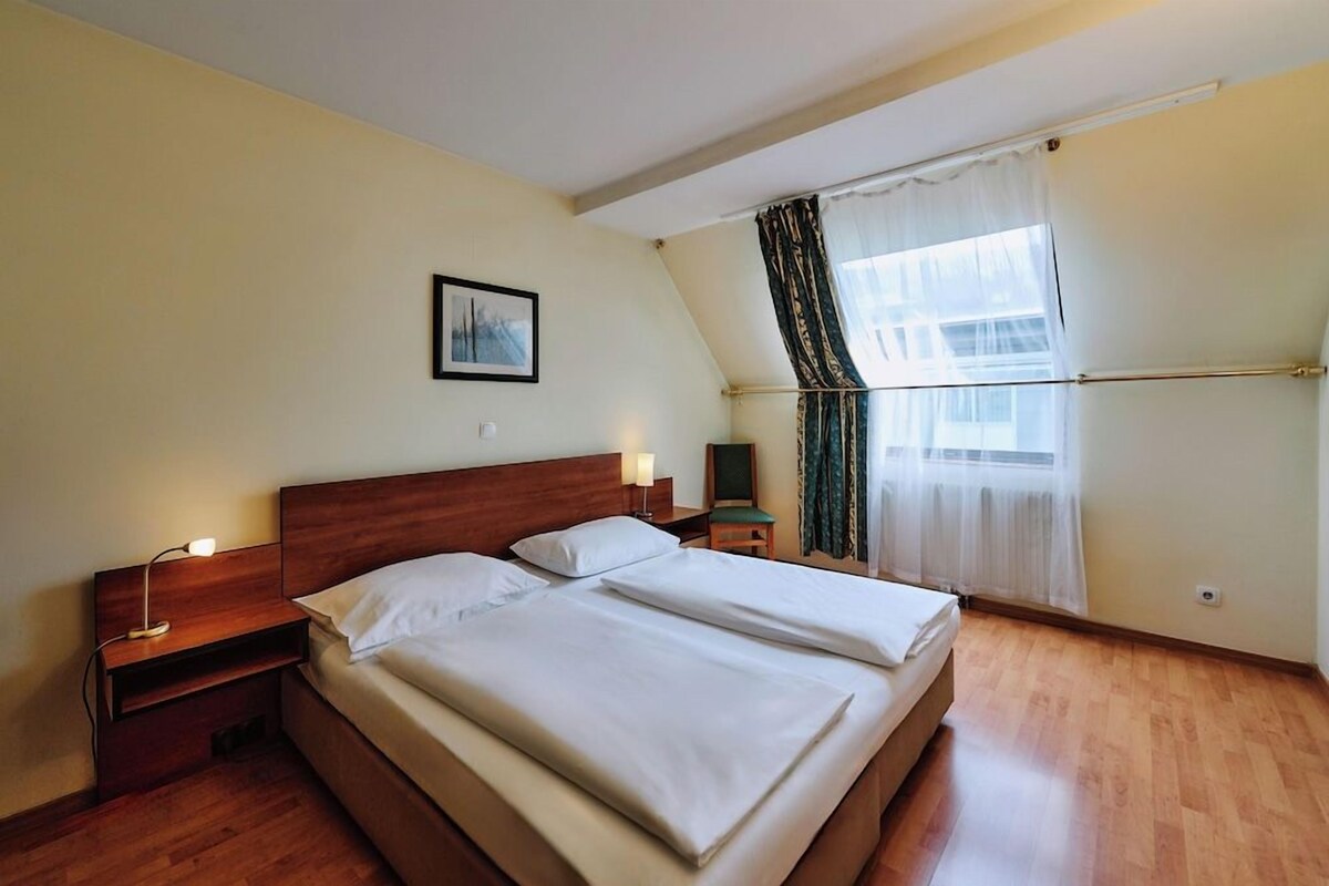 Ein Schlafzimmer mit einem Doppelbett, Nachttischen, einer Lampe, einem gerahmten Bild an der Wand und einem Fenster mit Vorhängen.