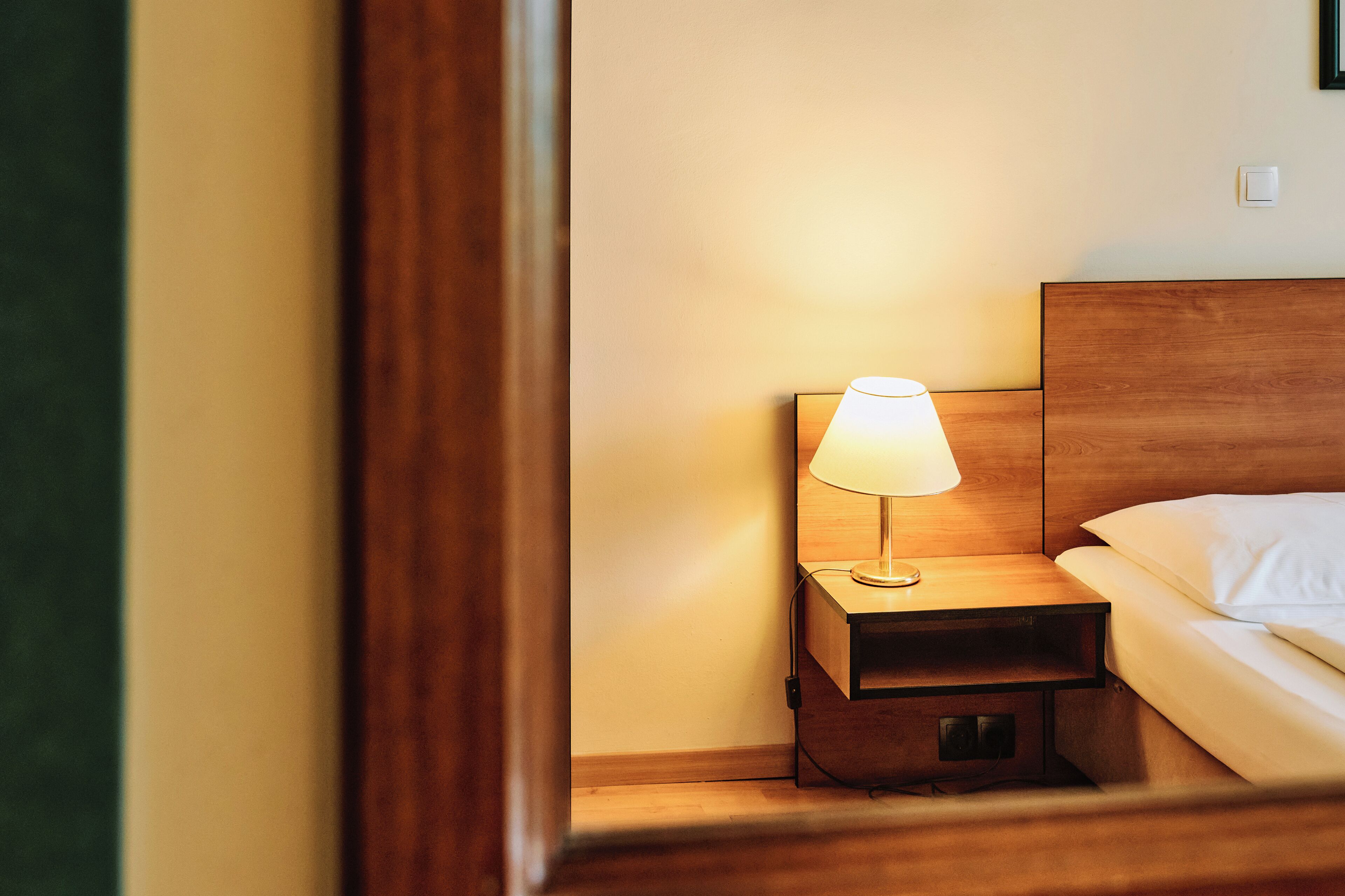 Una cama bien hecha con cabecera de madera, una mesita de noche con lámpara y un enchufe en la pared.