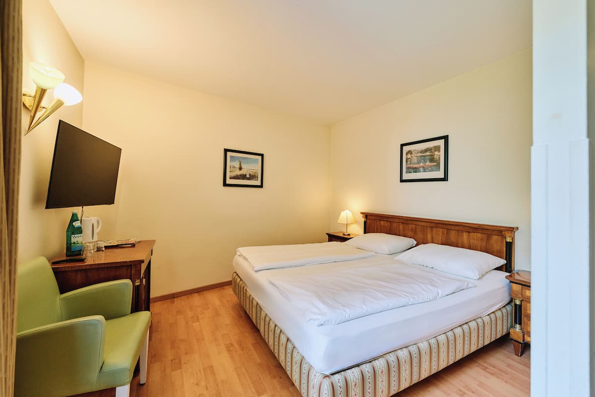 A hotel room with a double bed, a green chair, a desk with a lamp, and two framed pictures on the wall.