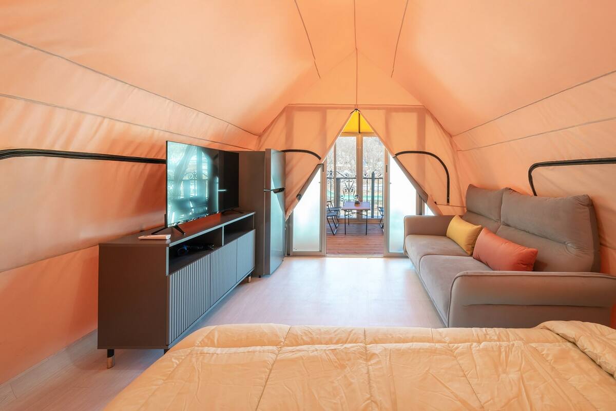 Una habitación tipo tienda de campaña acogedora y bien iluminada, con cama, sofá, televisión y zona de comedor con mesa y sillas.