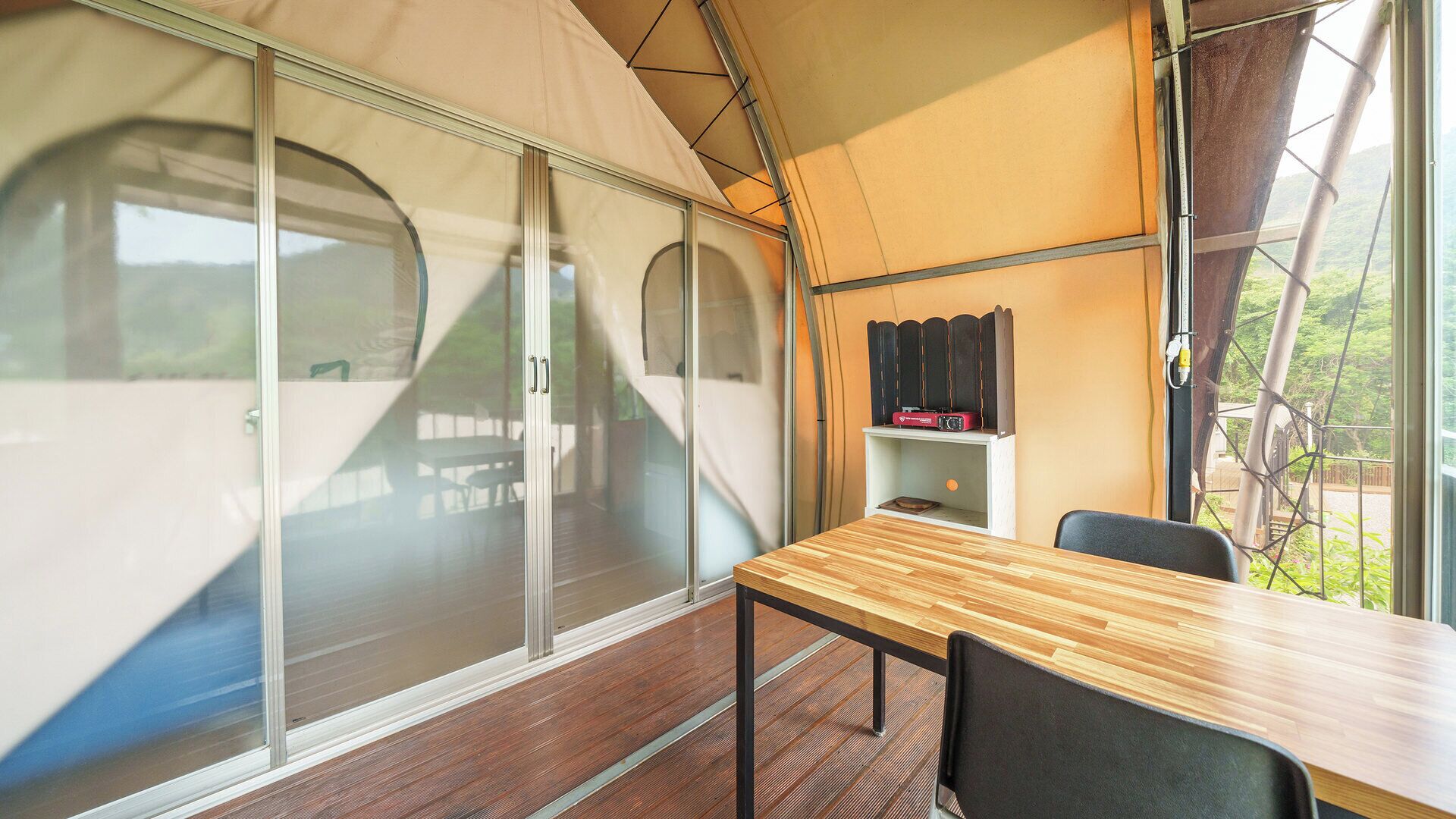A tent with a wooden table and chairs, large windows, and a shelf with a radio.