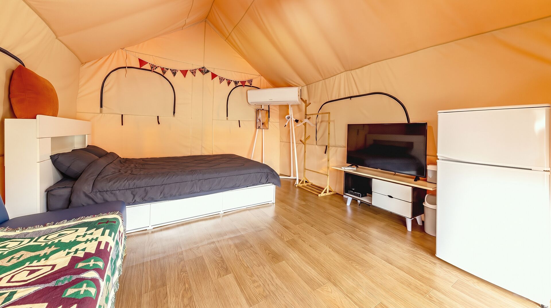 A tented room with a bed, a television, a refrigerator, and a portable air conditioning unit.