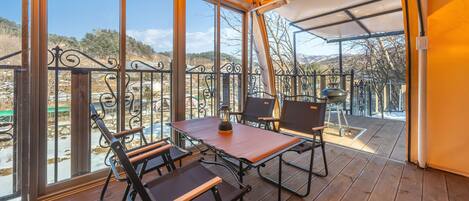 A covered outdoor seating area with a table and chairs, a grill, and a view of the surrounding landscape.