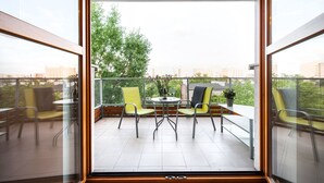 Un balcón con una mesa y sillas, una planta en maceta, y vistas a zonas verdes y edificios.