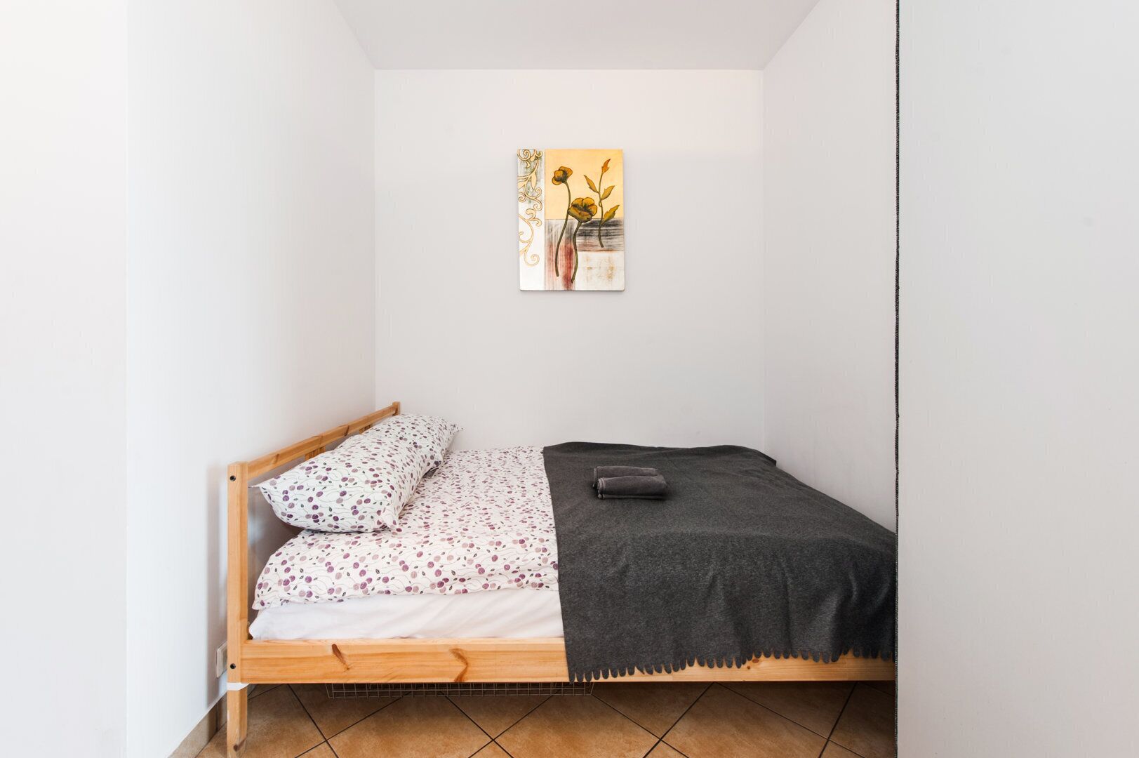 Ein kleines Zimmer mit einem Einzelbett, einem hölzernen Bettgestell, floralem Bettbezug, einer grauen Decke und einem gerahmten Kunstwerk an der Wand.