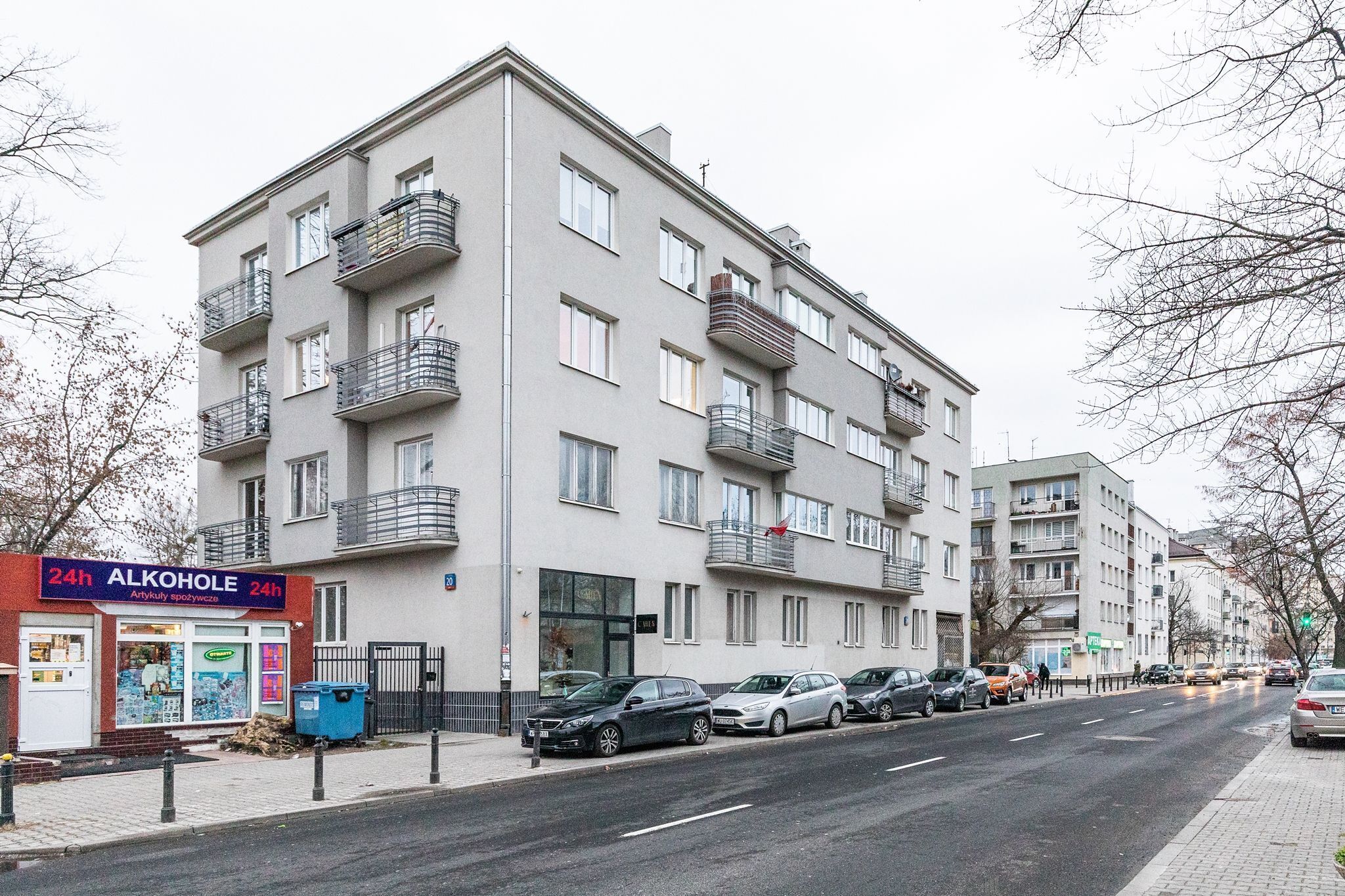 Ein mehrstöckiges Wohngebäude mit Balkonen, ein Getränkehandel mit rund um die Uhr geöffnet, und parkende Autos am Straßenrand.