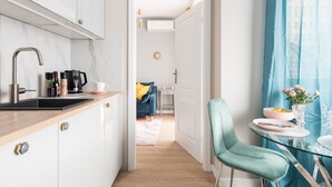 Una cocina moderna con encimera negra, armarios blancos y grifo de acero inoxidable. Hay un comedor con una silla azul, una mesa de vidrio y un florero con flores.