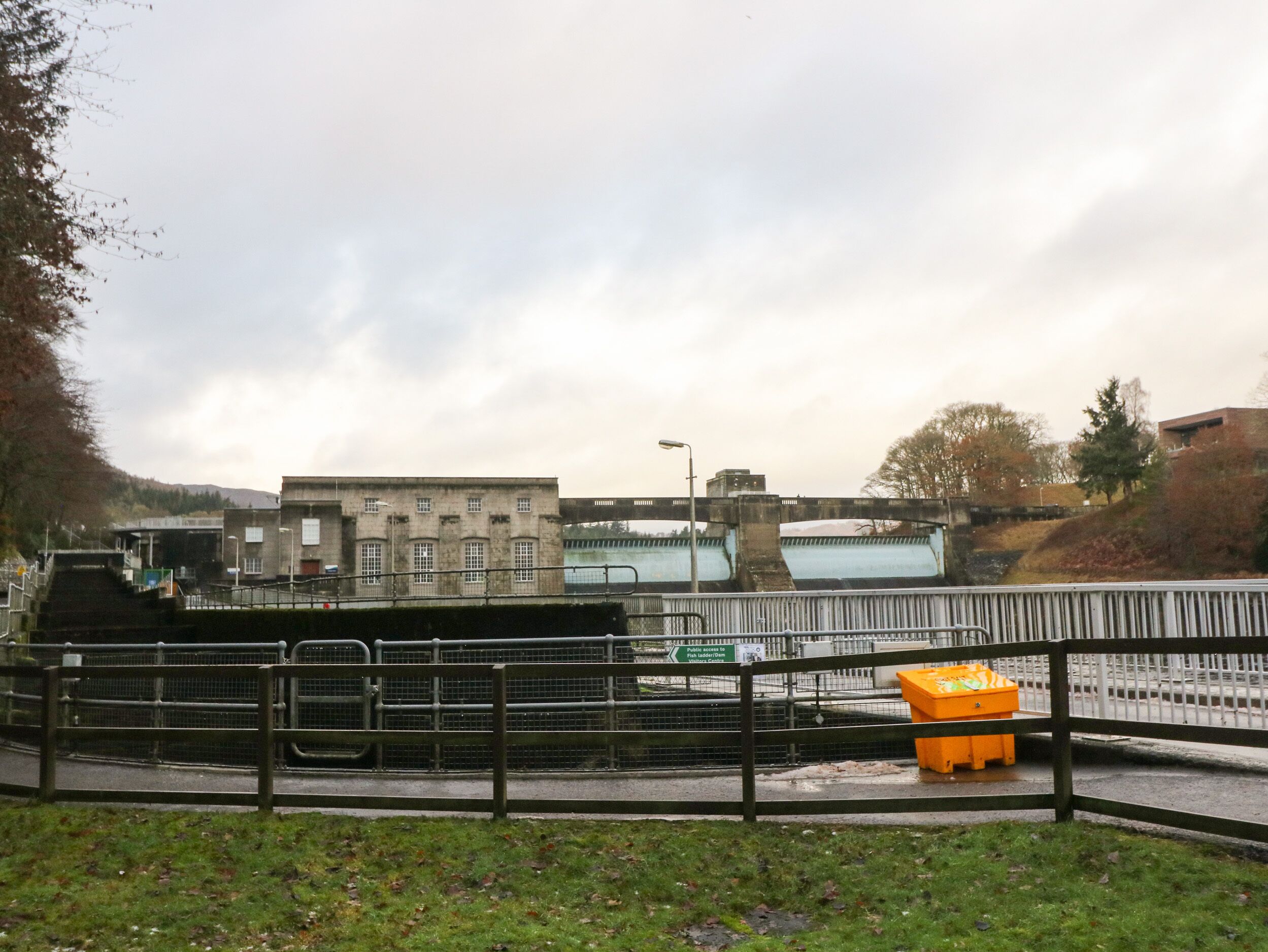 Een brug met een metalen hekwerk, een grasveld op de voorgrond en een gebouw met een bord achterin.