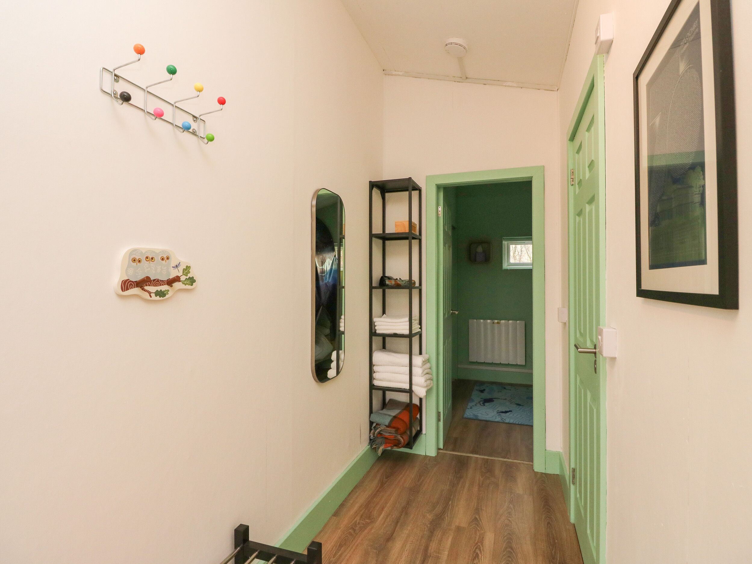 Un pasillo con piso de madera, una puerta verde, un espejo, una estantería con toallas e iluminación empotrada en la pared.