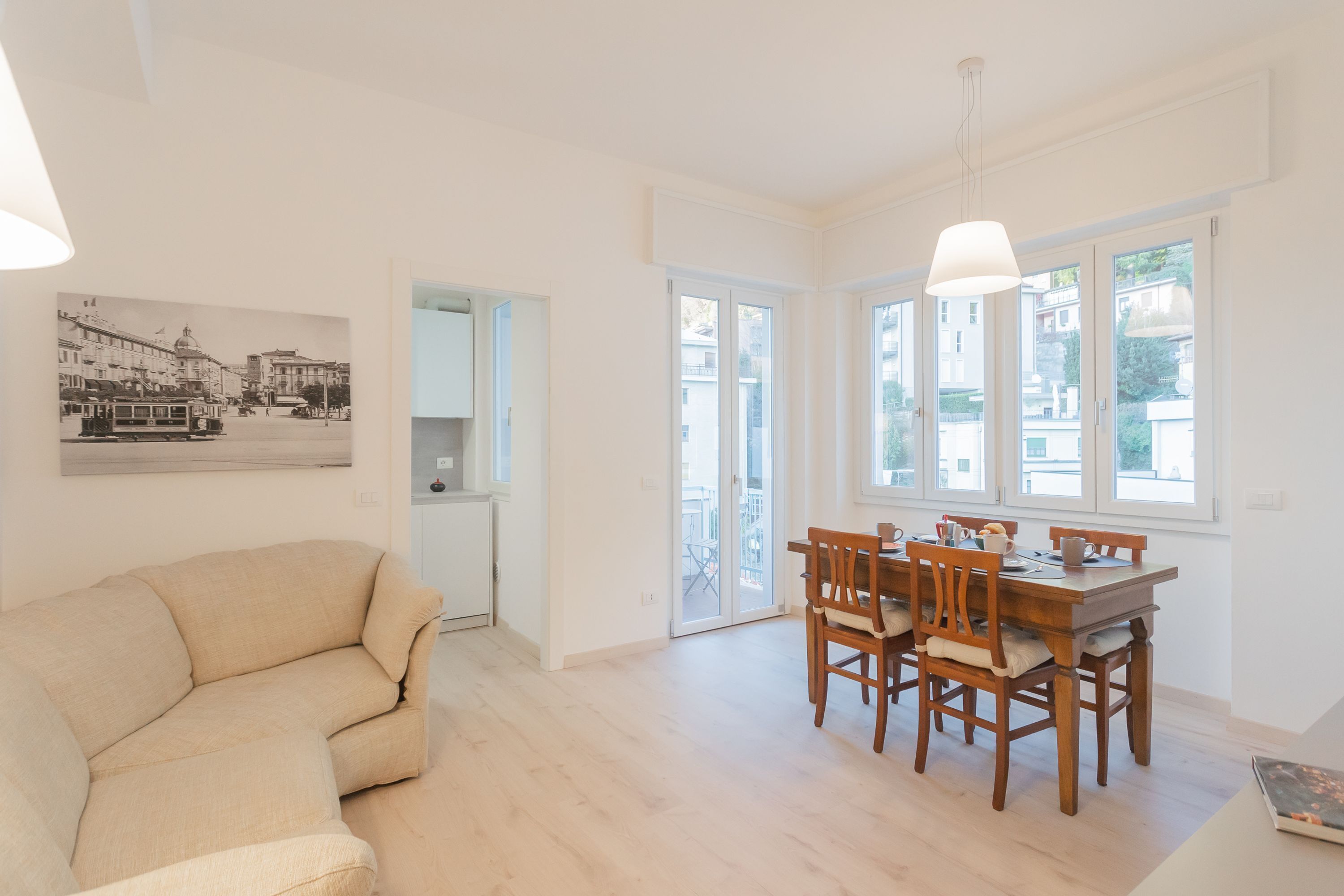 A spacious living room with a beige sofa, a wooden dining table with chairs, and a large window with a view of the street.
