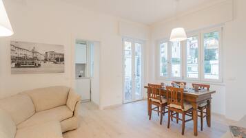 A spacious living room with a beige sofa, a wooden dining table with chairs, and a large window with a view of the street.