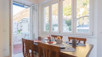 A dining area with a wooden table and chairs, a window with a view of buildings and hills, and a door leading to an outdoor space with a table and chairs.