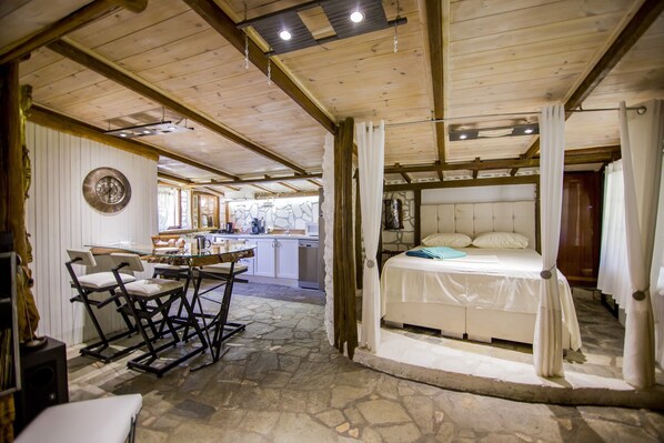 Una habitación acogedora con una cama, un comedor con sillas, una cocina al fondo y suelo de piedra.