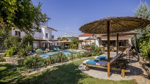 Un espace piscine avec des chaises longues et un parasol en chaume, entouré de verdure et de bâtiments.