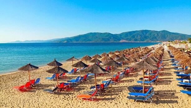 Une plage bordée de rangées de chaises longues et d’ombrelles en chaume, une mer d’un bleu limpide et des montagnes en arrière-plan.
