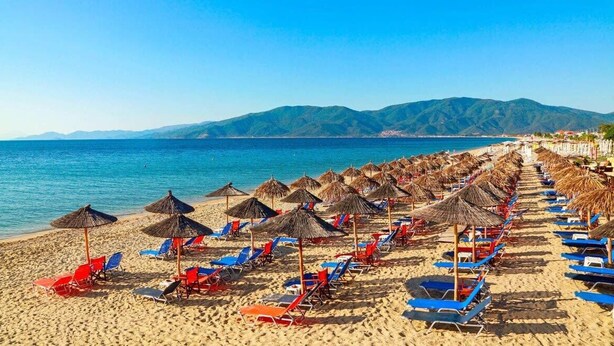 Une plage bordée de rangées de chaises longues et d’ombrelles en chaume, une mer d’un bleu limpide et des montagnes en arrière-plan.