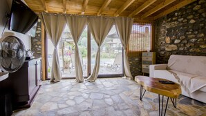 Une pièce avec un sol en pierre, un plafond en bois, un canapé blanc, une table basse en bois, un téléviseur fixé au mur et des rideaux aux fenêtres.