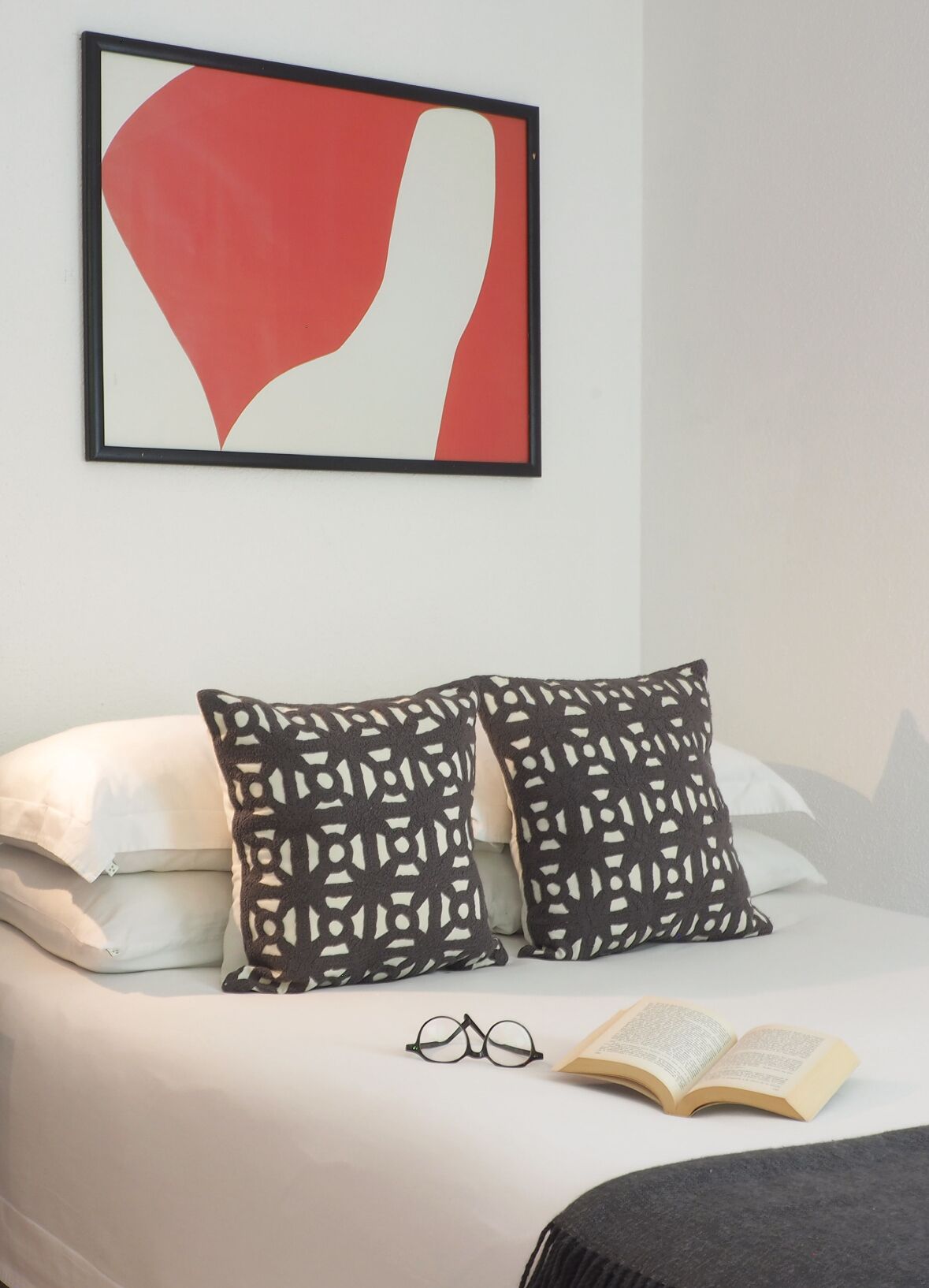 A neatly made bed with white bedding, two patterned pillows, a framed abstract artwork, and an open book with glasses beside it.