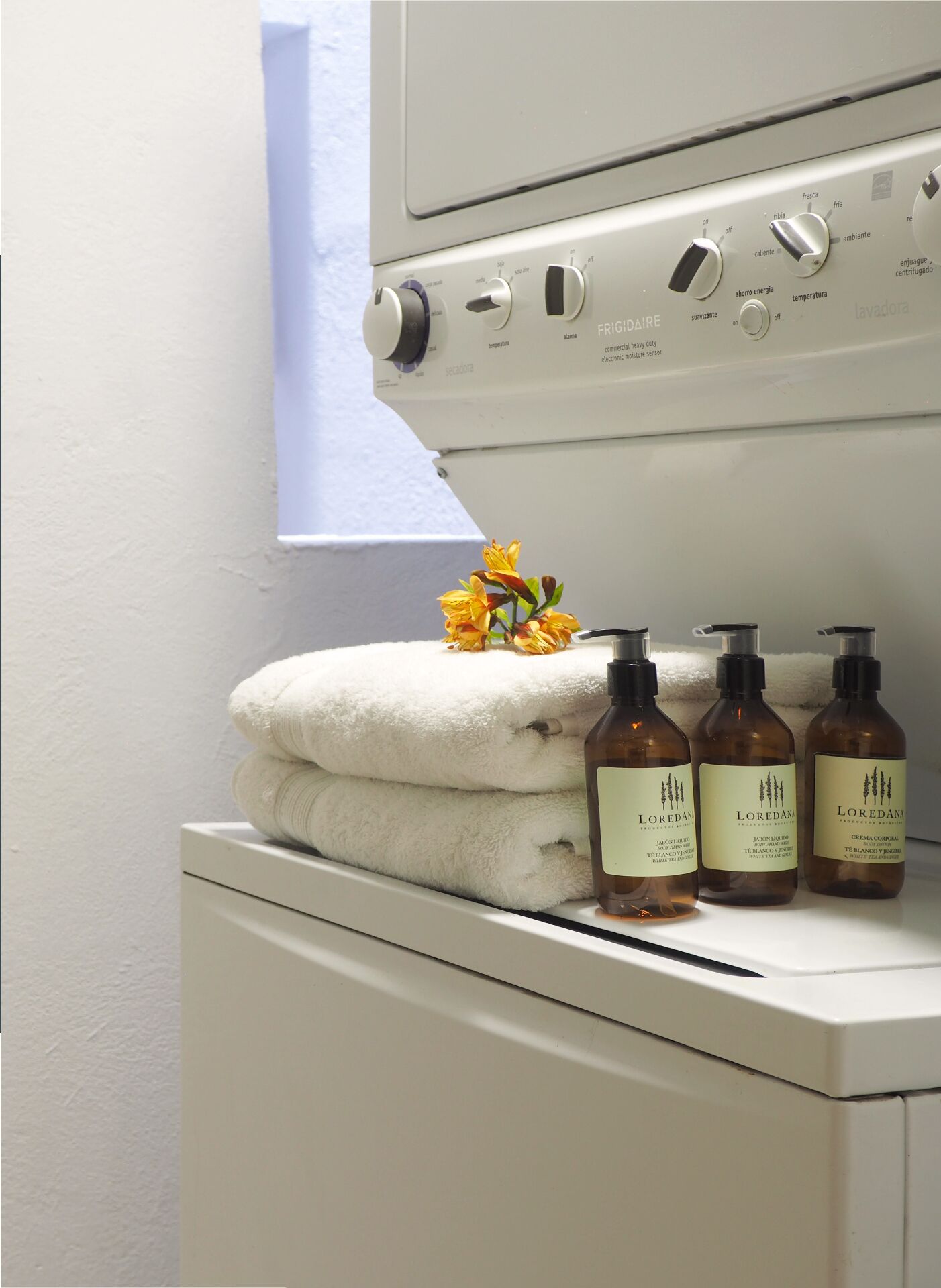 A white washing machine with control knobs and a window above it. Three bottles of Loredana cream are placed on top of a folded white towel.