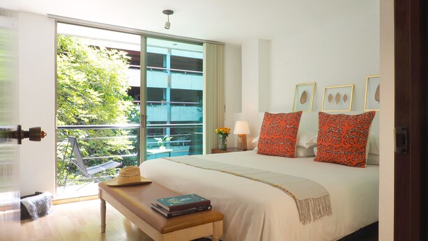 A bedroom with a large bed, a bench, a bedside table with a book, and a view of a balcony with greenery. - Comfort Reforma By The Local Way (Mexico City)