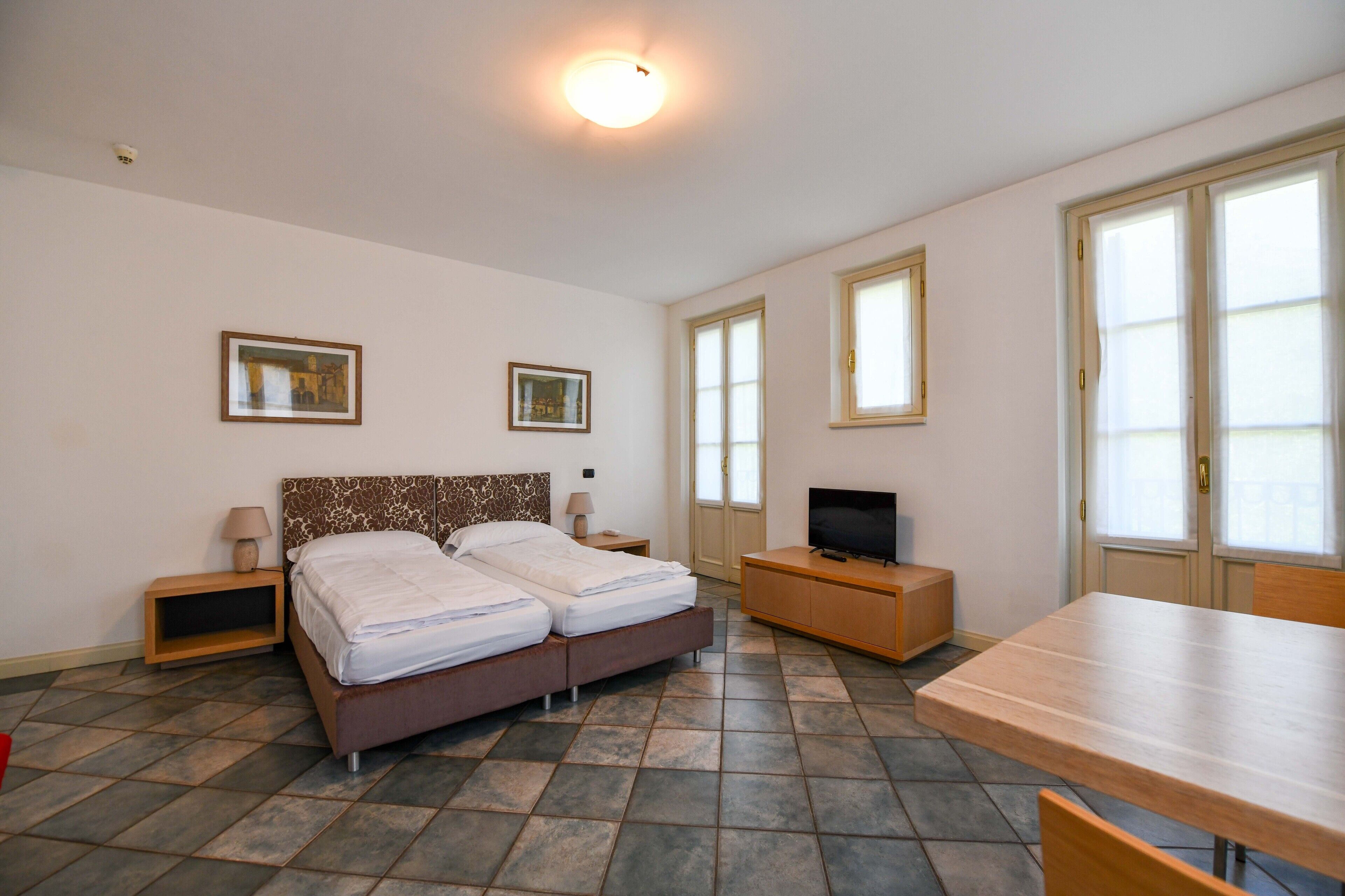 A bedroom with a double bed, a TV, a wooden table, and two windows.