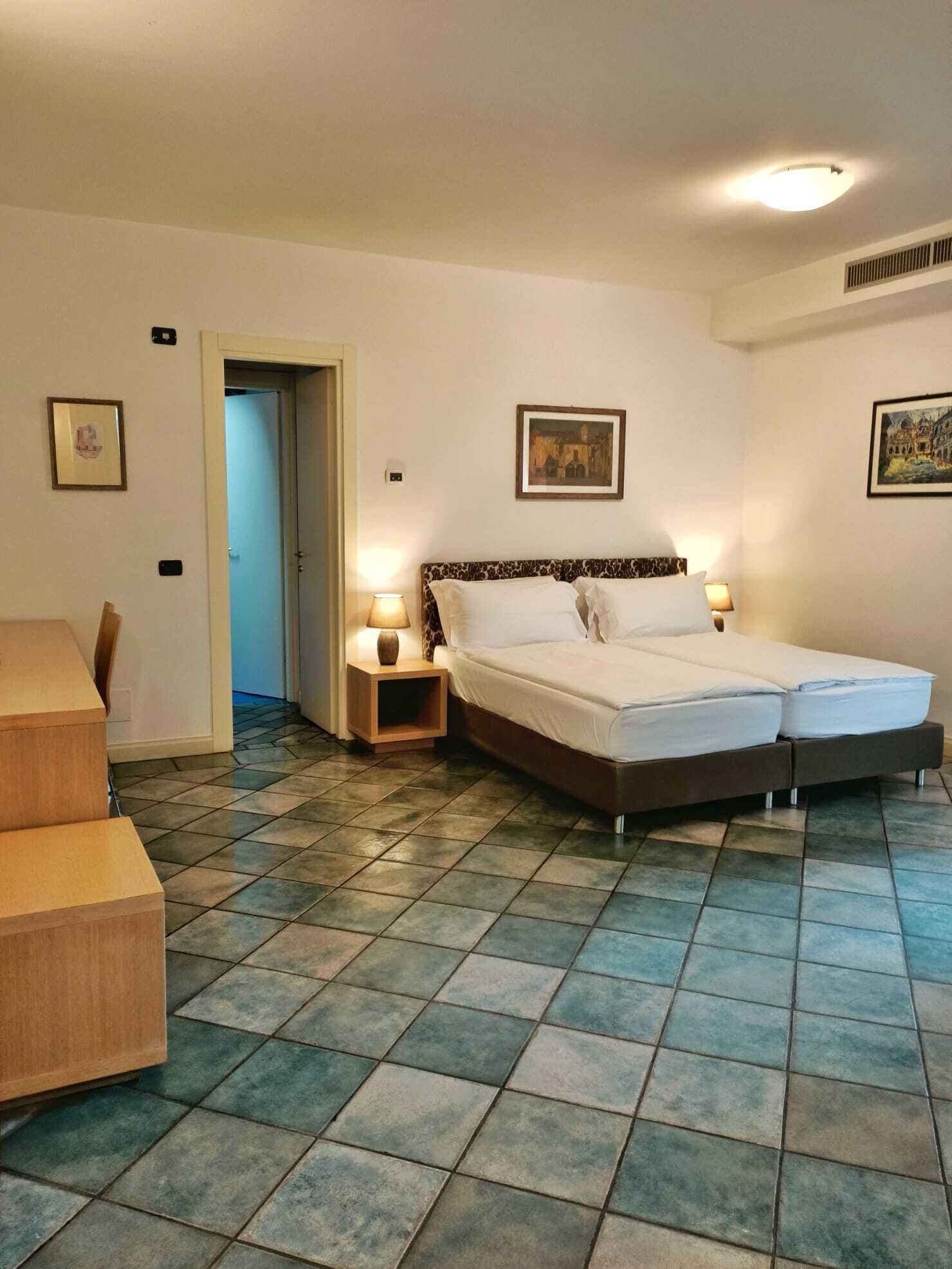 A hotel room with a double bed, two bedside tables with lamps, a wooden chair, and a tiled floor.