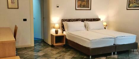 A hotel room with a double bed, two bedside tables with lamps, a wooden chair, and a tiled floor.