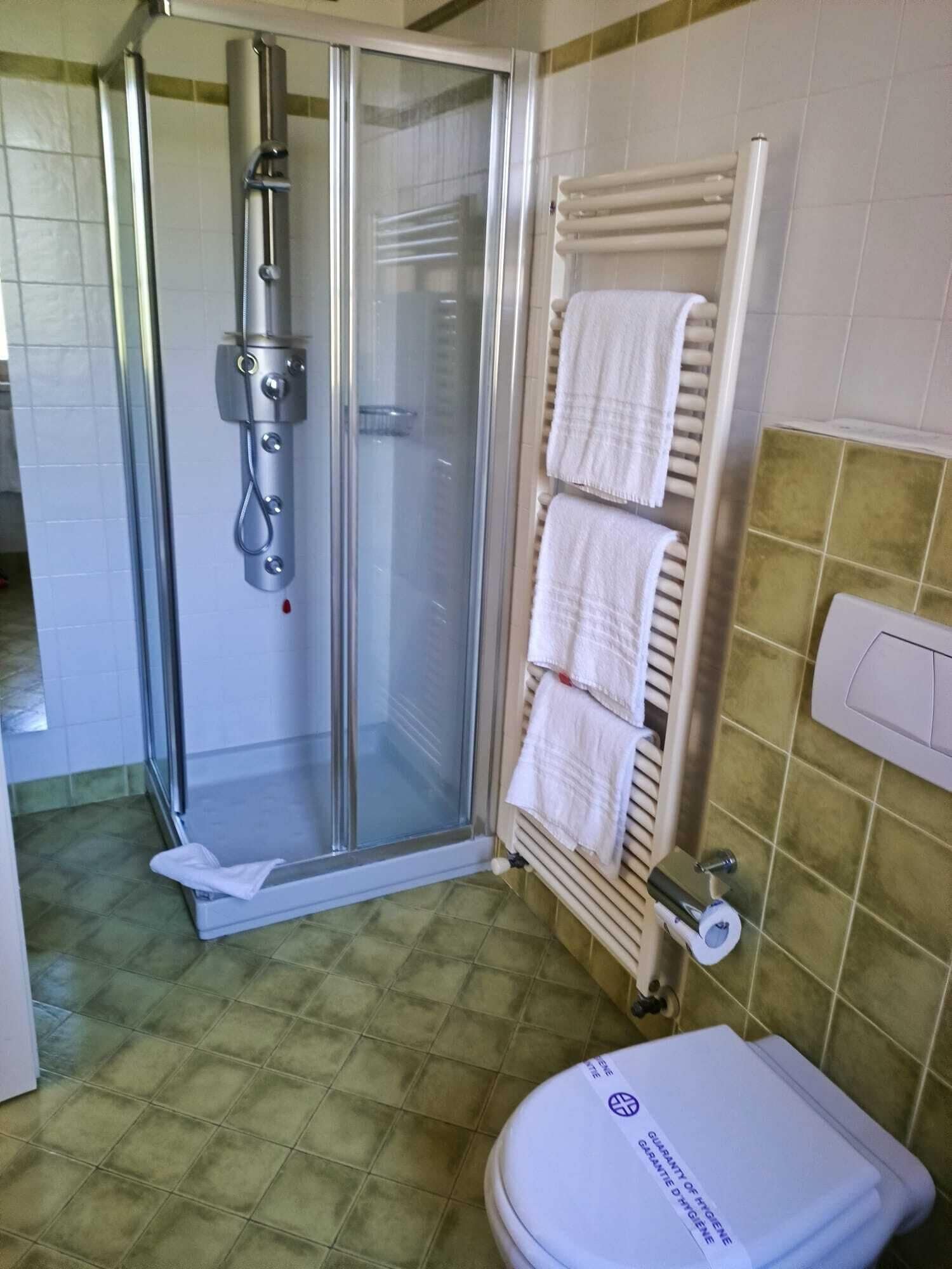 Un baño con cabina de ducha de vidrio, un perchero con varias toallas y un inodoro con etiqueta de garantía de higiene.
