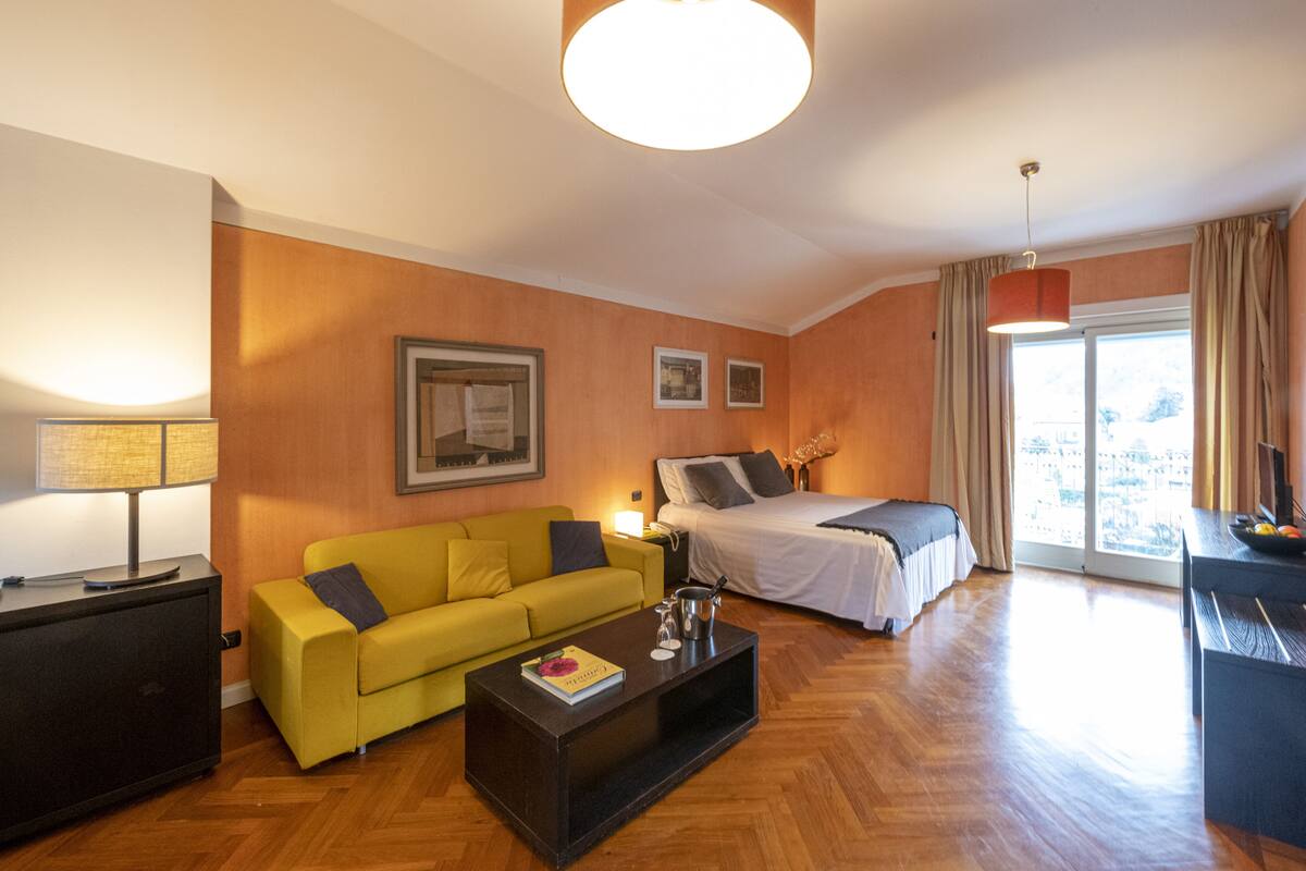 Een hotelkamer met een gele bank, een bed, een bureau en een groot raam met gordijnen.