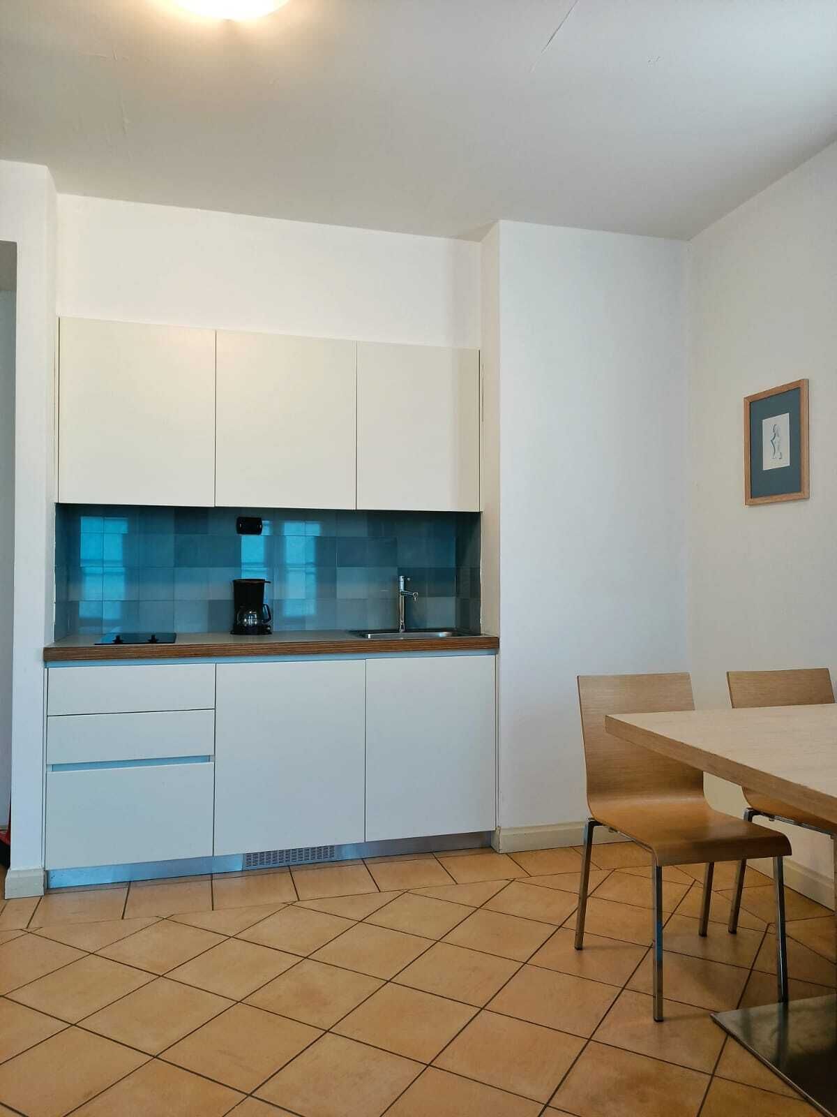 Eine moderne Küche mit weißen Schränken, einer blauen Spritzschutzwand und einer Kaffeemaschine. Es ist ein Esstisch mit Stühlen und ein gerahmtes Bild an der Wand zu sehen.