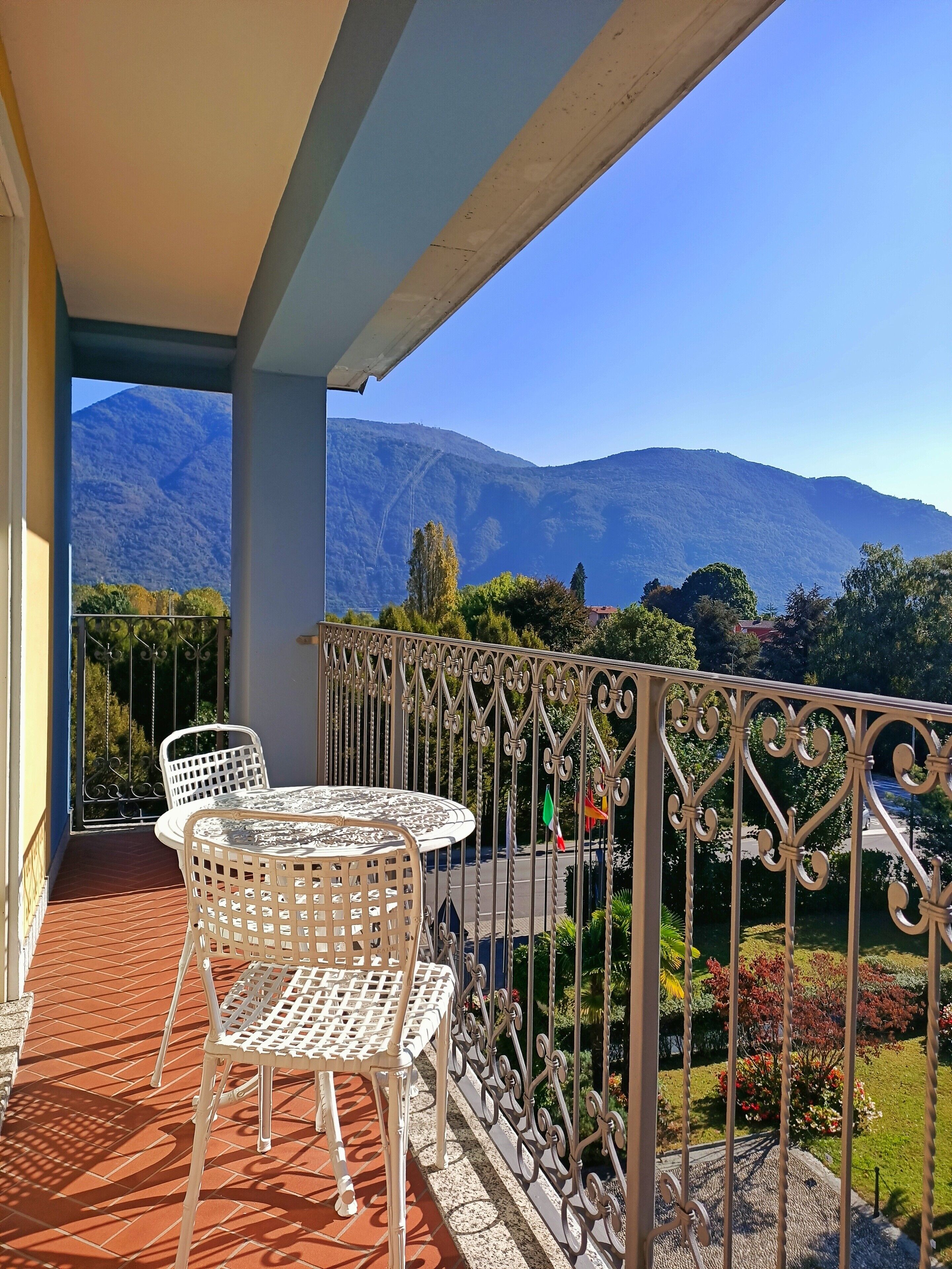 A balcony with a table and chairs, a view of mountains, and a garden.