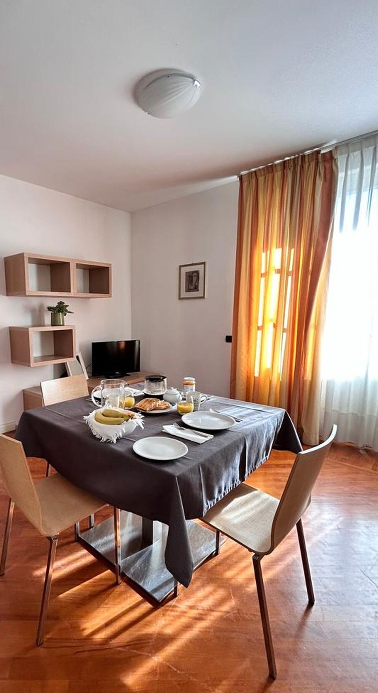 A dining area with a table set for four, a television on the wall, and a window with curtains.