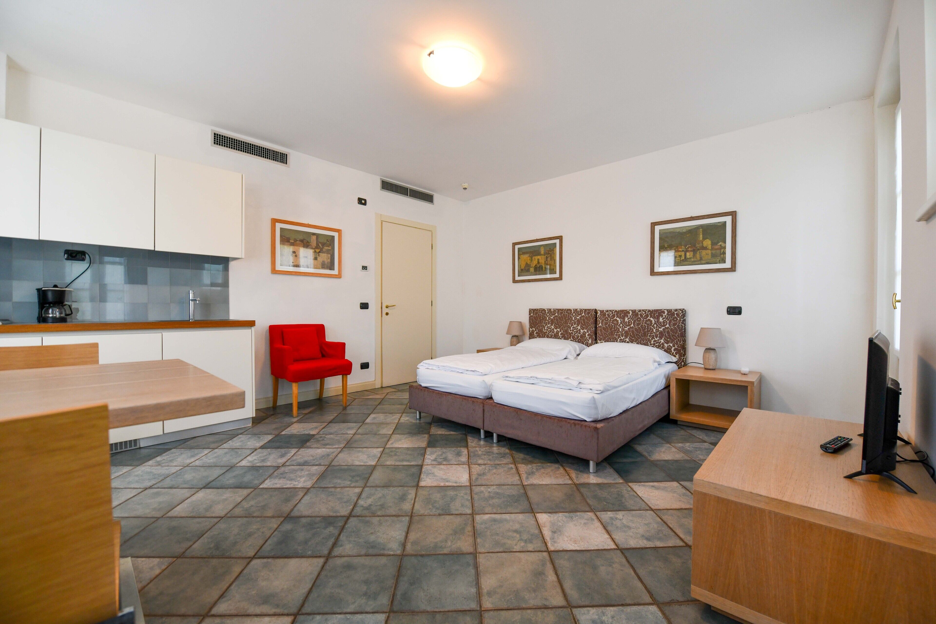 A hotel room with a bed, a desk, a chair, a TV, and a small kitchen area.
