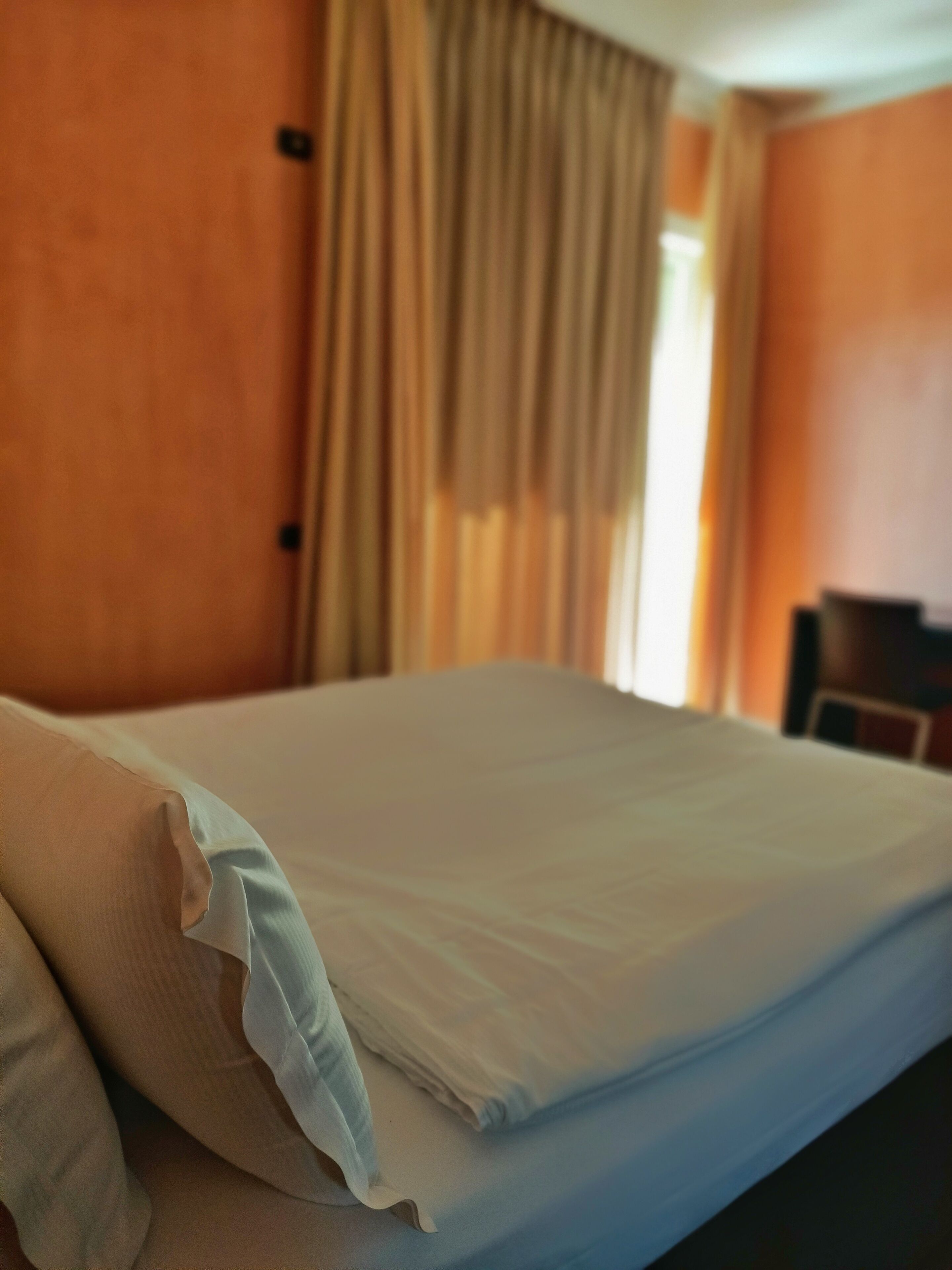A neatly made bed with pillows, a window with curtains, and a chair in a room with orange walls.