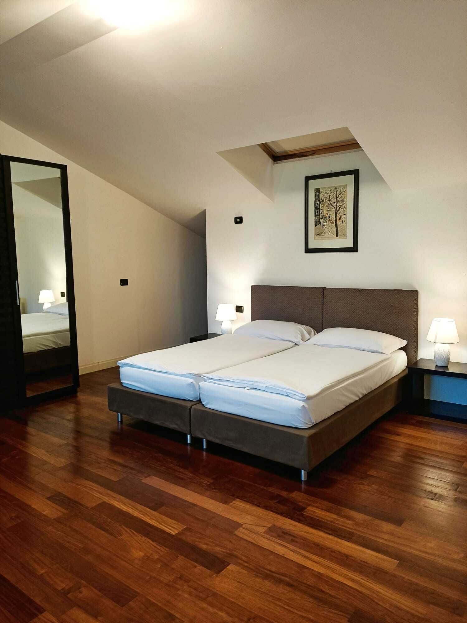 Une chambre à coucher avec un lit double, un plancher en bois, un luminaire au plafond, un tableau encadré et une table de chevet avec une lampe.
