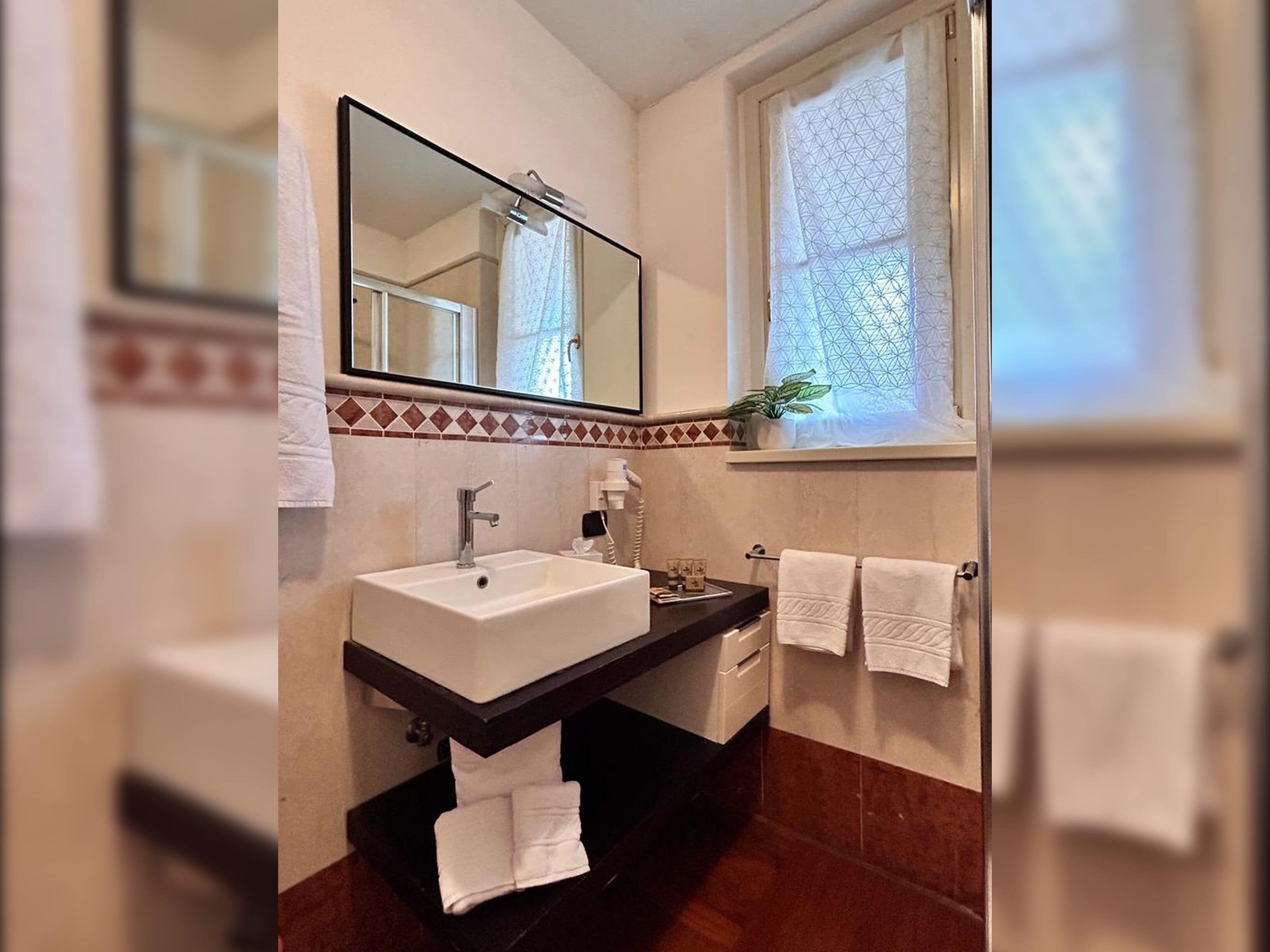 Een badkamer met een witte wasbak, een zwart aanrecht, een spiegel en een raam met een geometrisch gordijn.