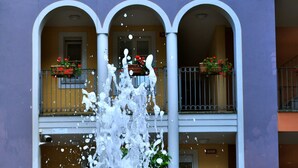 Une fontaine d’eau se trouve devant un immeuble doté de balcons et orné de plantes en pot.