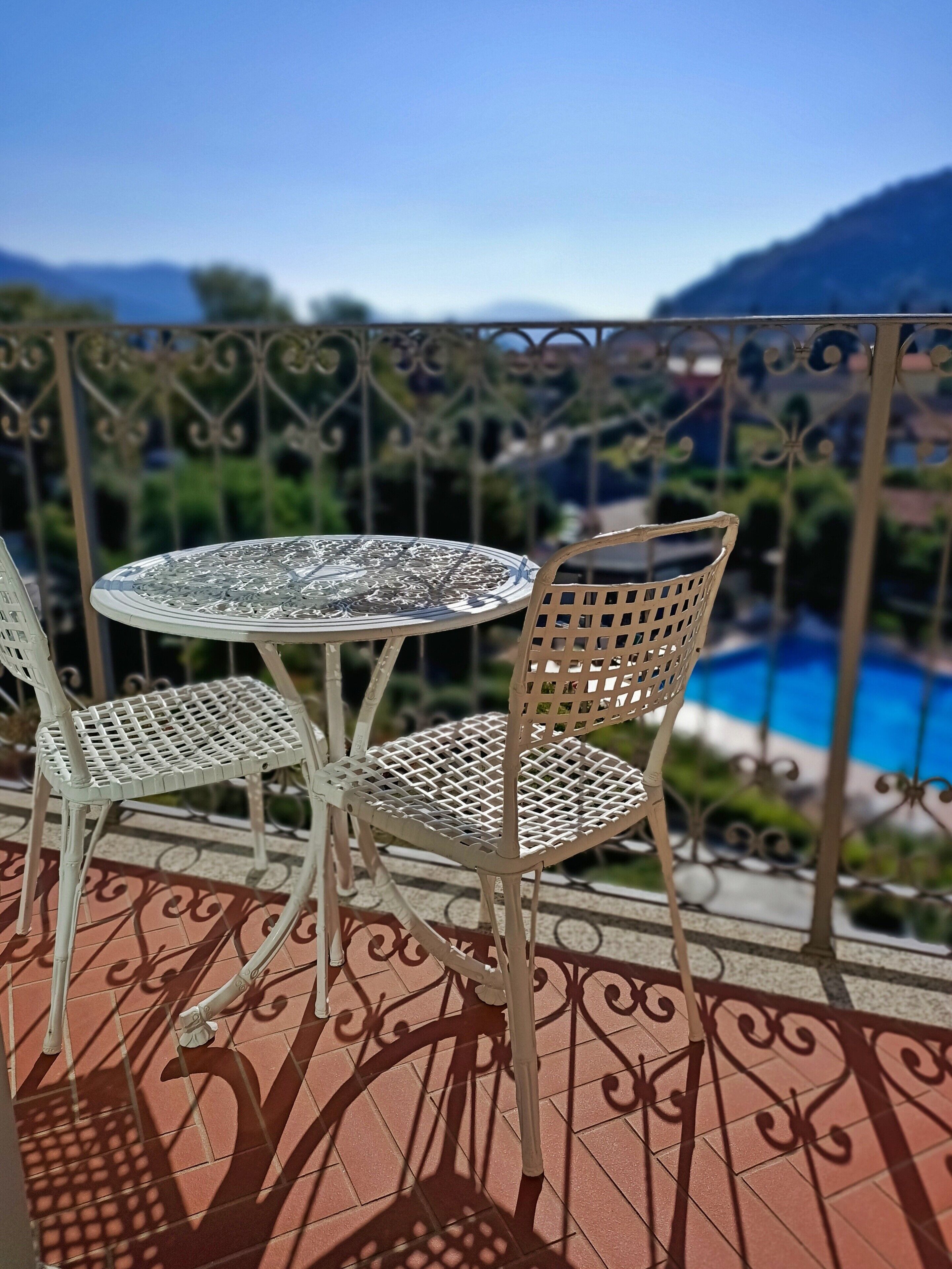 A balcony with a table and two chairs, overlooking a swimming pool and greenery.