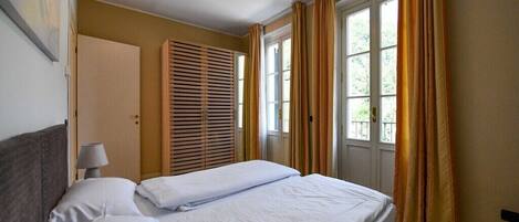 Una habitación de hotel con cama, mesita de noche, lámpara, ventana con cortinas y puerta.
