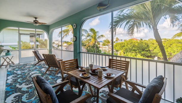 A balcony with wooden furniture, a patterned rug, and a view of palm trees and buildings. - Reel Knotty ~ Pool Home, 35' Dock, Walk to the Beach, Bikes & Kayaks! (Marathon)