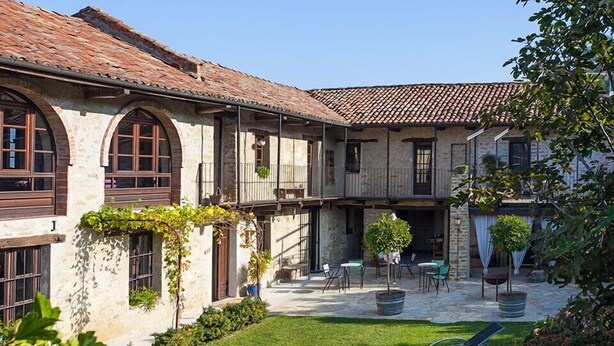 A stone building with arched windows and a tiled roof, featuring a courtyard with outdoor seating and a lawn. - Cascina IL SOGNO  (Cossano Belbo)