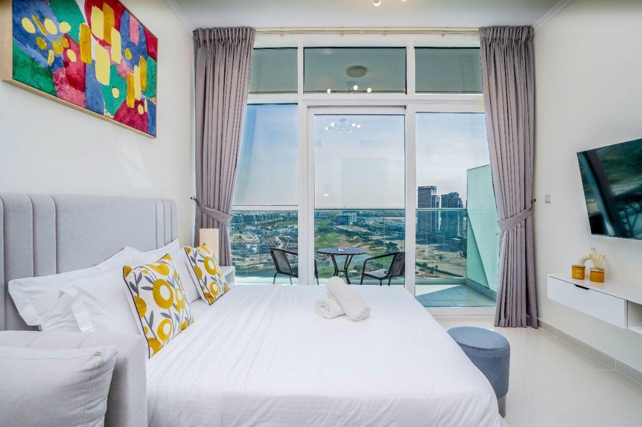 A modern hotel room with a large bed, a balcony with a city view, and a flat-screen TV on the wall.
