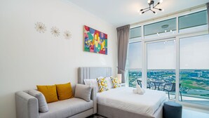 Un dormitorio moderno con un sofá gris, una cama con ropa de cama blanca, un cuadro abstracto colorido y un ventanal con vistas a la ciudad.