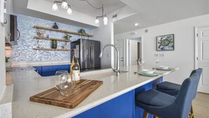 A modern kitchen with a blue accent wall, stainless steel appliances, and a wooden island with bar seating. - Gulf Front 3/3 Resort w/Pool & 3 King Suites! Chair/umbrella set-up included! (Navarre)