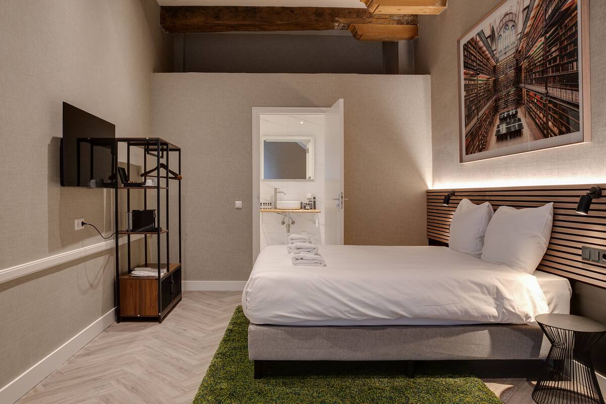 A modern hotel room with a large bed, a green rug, a wooden headboard, a wall-mounted TV, and a bookshelf.