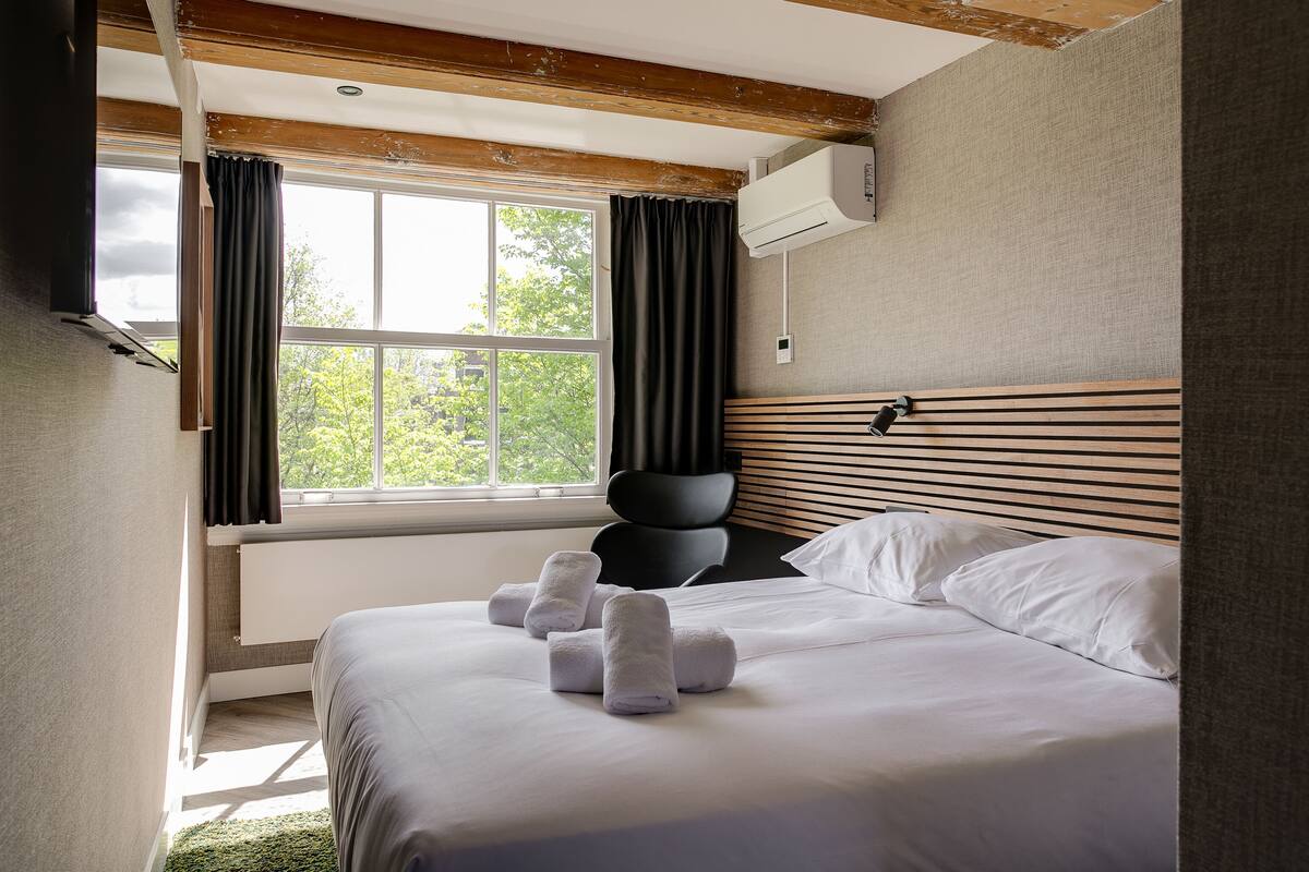 A hotel room with a large bed, a window with curtains, a ceiling fan, and a wall-mounted air conditioner.