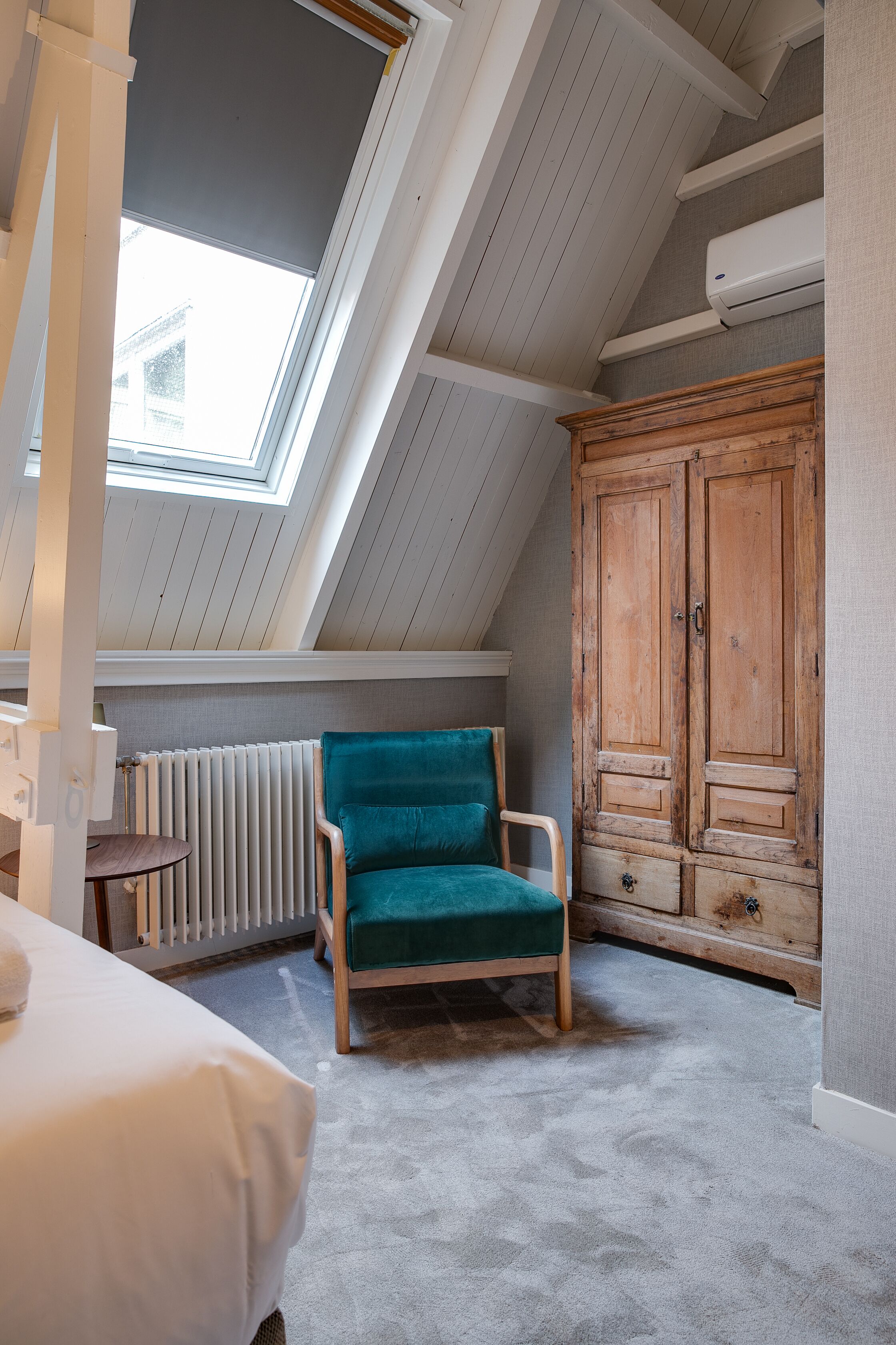 A room with a sloped ceiling, a wooden wardrobe, a teal armchair, and a radiator.